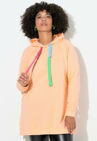 Mujer con cabello rizado que lleva una sudadera melocotón con cordones rosas, azules y verdes, aretes de aro verdes y pantalones de cuero negros.