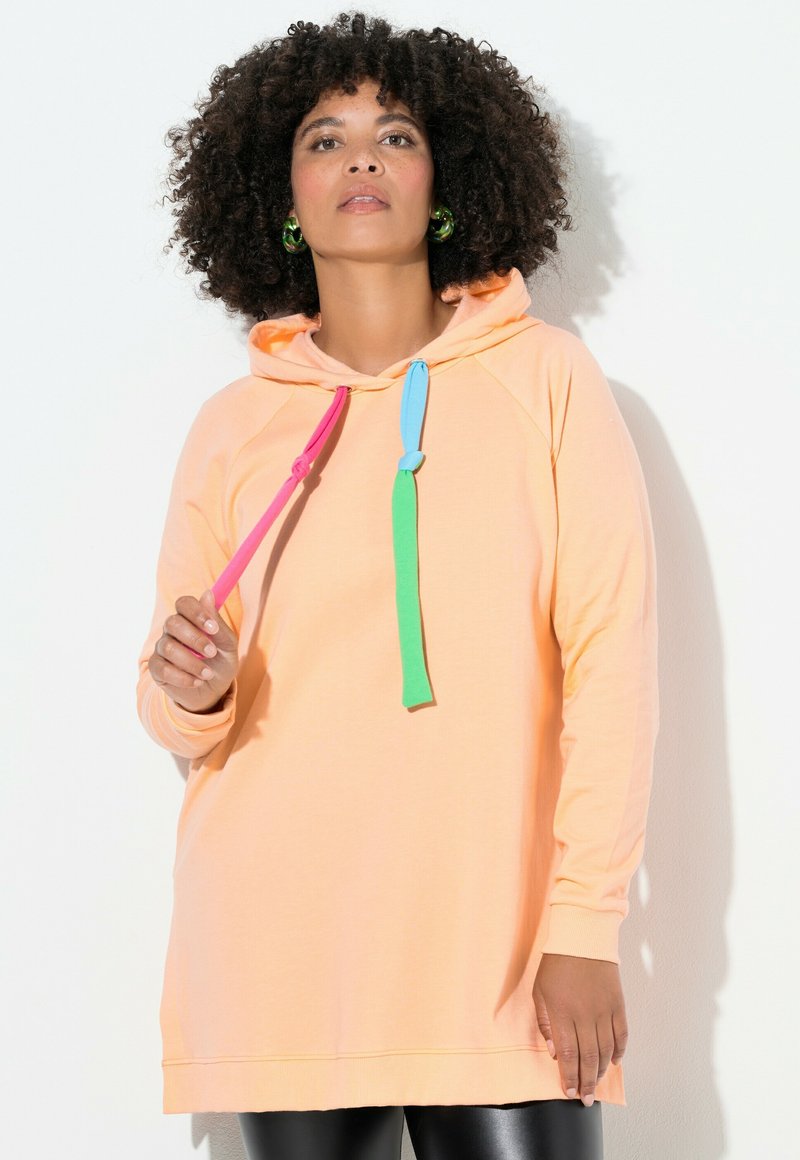 Mujer con cabello rizado que lleva una sudadera melocotón con cordones rosas, azules y verdes, aretes de aro verdes y pantalones de cuero negros.