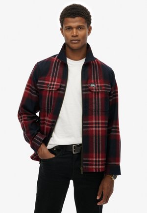 Chaqueta de franela a cuadros en rojo y azul marino con dos bolsillos en el pecho, cierre de cremallera y cuello plano, diseñada con una textura suave y un corte casual.