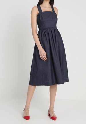 Vrouw in een mouwloze donkerblauwe midi-jurk met een vierkante halslijn en beige schoenen met rode puntige neuzen en blokhakken.