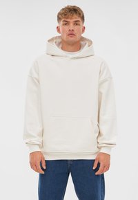 Sudadera de color crema hecha de un material suave, con un gran bolsillo frontal, capucha con cordones y un corte holgado, combinada con jeans azules.