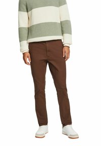 Uomo che indossa pantaloni slim fit marroni, sneakers bianche e un maglione a maniche lunghe a righe verdi e bianche, in piedi su uno sfondo bianco.