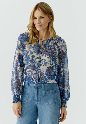 Vrouw draagt een doorschijnende blouse met lange mouwen en blauwe paisley-patronen, gecombineerd met een hoge taille blauwe spijkerbroek, staand tegen een effen lichte achtergrond.