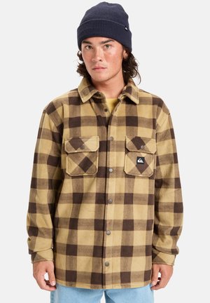 Brown and tan plaid flannel shirt with two chest pockets and snap buttons. Worn over a yellow shirt, paired with light blue jeans.