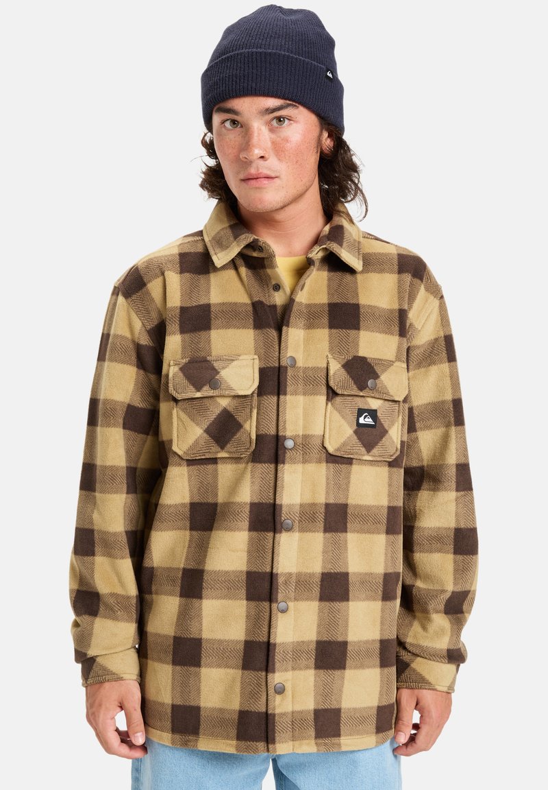 Braun und beige karierter Flanellhemd mit zwei Brusttaschen und Druckknöpfen. Über einem gelben Hemd getragen, kombiniert mit hellblauen Jeans.