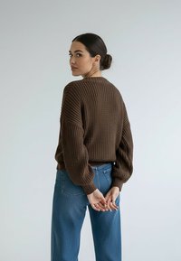 Brązowy sweter dziany z teksturowanym wzorem, o luźnym kroju i balonowych rękawach, zestawiony z wysokim stanem niebieskich dżinsów.