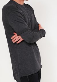 Mann in dunkelgrauem Langarmshirt und schwarzer Hose, der mit verschränkten Armen vor einem schlichten weißen Hintergrund steht.