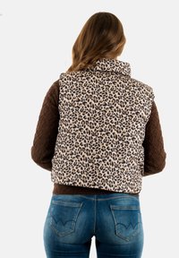 Maglioncino marrone trapuntato a maniche lunghe sotto un gilet beige con stampa leopardata e colletto alto, indossato con jeans in denim blu con tasche posteriori.