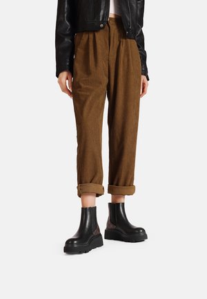 Pantaloni a righe marroni a gamba affusolata con risvolti, abbinati a stivaletti neri alla caviglia con pannelli testurizzati e suole spesse.