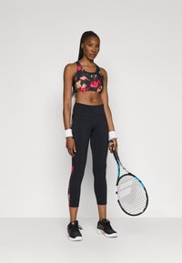 Fekete alapon színes virágmintás sportmelltartó; magas derekú fekete leggings virágmotívummal az oldalán; teniszütővel párosítva.