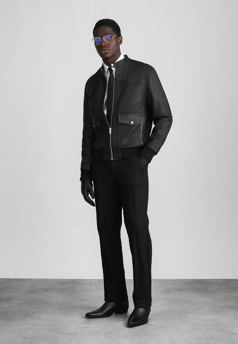 Black leather bomber jacket with two front pockets, paired with textured black pants and sleek black ankle boots, wearing gloves and glasses.