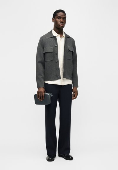 Άνδρας με σκούρο γκρι σακάκι με κουμπιά πάνω από λευκό πουκάμισο και μαύρο παντελόνι, κρατώντας μια μικρή γκρι τσάντα στο δεξί του χέρι.