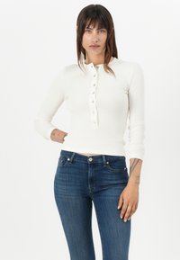Haut à manches longues blanc avec un col henley et cinq boutons, associé à un jean bleu foncé, silhouette ajustée et texture côtelée.