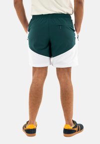 Calções de atletismo verdes e brancos com uma cintura elástica, bolsos laterais e detalhes em painel de contraste. Usados com ténis pretos e amarelos.