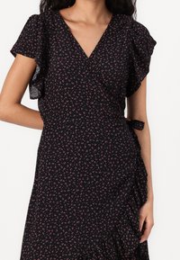 Abito avvolgente nero con maniche a farfalla corte, caratterizzato da piccoli motivi floreali rosa e un dettaglio con cintura laterale. Il tessuto ha una consistenza morbida.