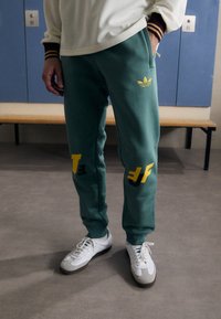 Green sweatpants with yellow accents and graphic text, featuring an embroidered logo. Paired with white sneakers.