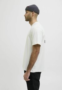 Cremefarvet bomulds t-shirt med korte ærmer, båret med mørke bukser og en grå ribbet beanie med rød detalje. Sideprofil.