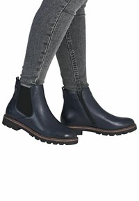 Piernas vestidas con jeans ajustados de color gris oscuro y botas de cuero azul marino hasta el tobillo con cremallera lateral y panel elástico sobre fondo blanco.