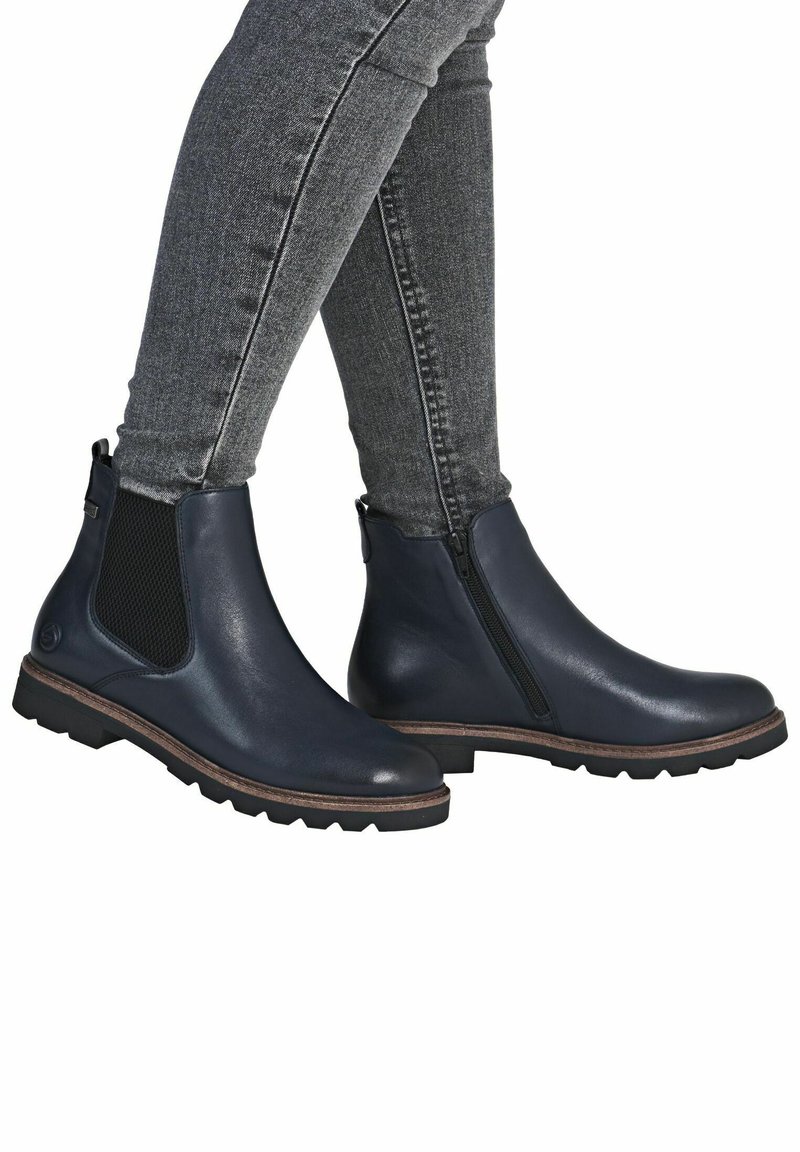 Piernas vestidas con jeans ajustados de color gris oscuro y botas de cuero azul marino hasta el tobillo con cremallera lateral y panel elástico sobre fondo blanco.