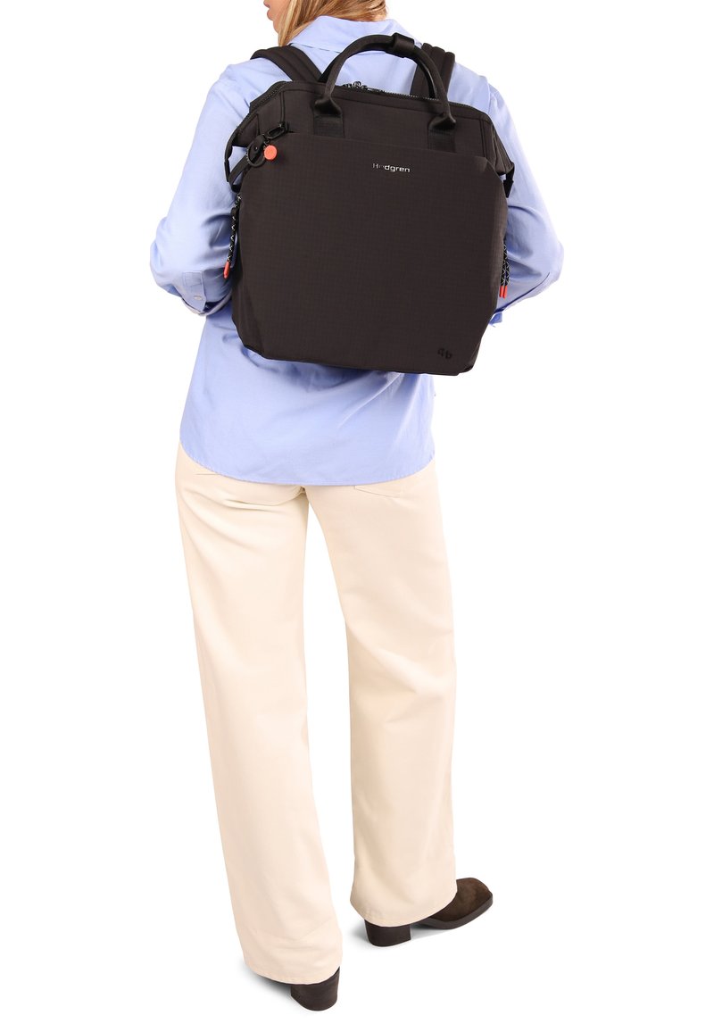 Personne portant une chemise bleu clair et un pantalon crème, portant un grand sac à dos noir avec des poignées dans le dos.