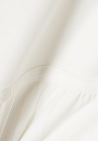 Gros plan d'un tissu blanc doux avec un motif floral en relief subtil le long d'une couture courbée, montrant la texture et les plis délicats.