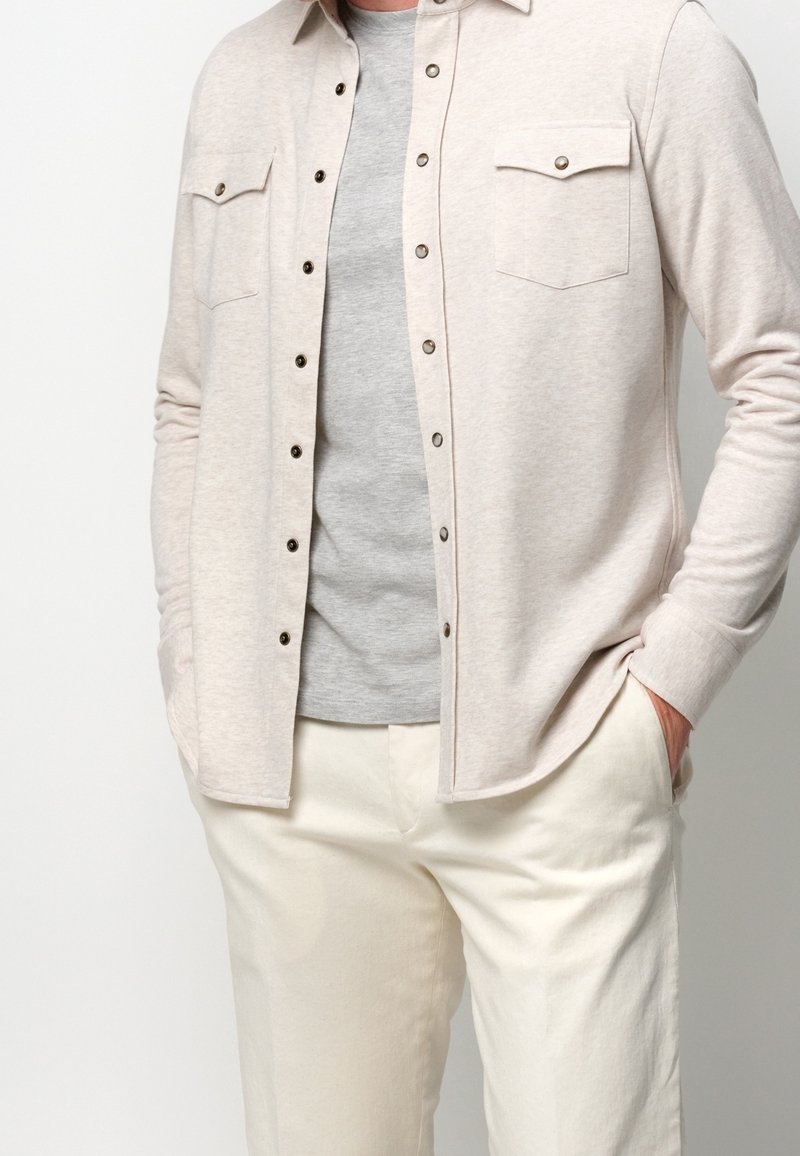 Homme portant une chemise beige clair à boutons par-dessus un t-shirt gris avec un pantalon couleur crème, mains dans les poches, sur un fond uni.