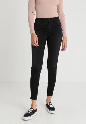 Woman wearing high-waisted black skinny jeans, beige long-sleeve fitted top, and black lace-up sneakers on a plain light background.