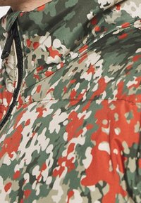 Gros plan sur une veste à motif camouflage vert, blanc, beige et rouge avec une fermeture éclair noire près du col.