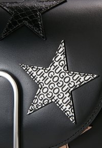 Detal detaliczny czarnej skórzanej torby z teksturowaną czarną aplikacją w kształcie gwiazdy oraz białą gwiazdą z powtarzającym się wzorem tekstowym "HUGO" w kolorze czarnym. Akcenty z srebrnym okuciem.