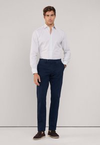 Lichtblauw geruit overhemd met een witte kraag, gecombineerd met marineblauwe broek en donkere instapschoenen, staande tegen een neutrale achtergrond.