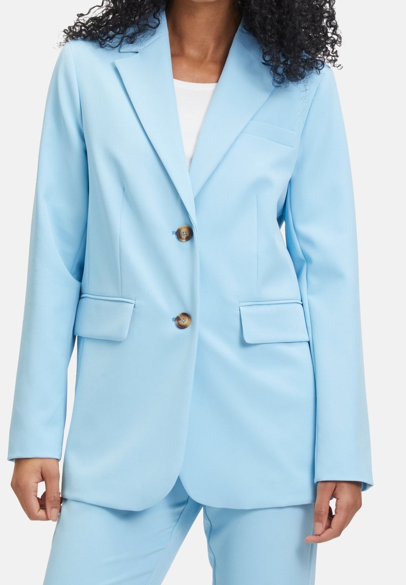 Femme portant un blazer bleu clair cintré à deux boutons et un pantalon assorti, un haut blanc en dessous, debout devant un fond uni.