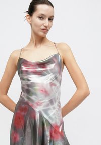 Robe nuisette en satin avec un décolleté drapé, présentant une base argentée et des motifs floraux abstraits rouges et verts, avec des bretelles fines et un ourlet évasé.