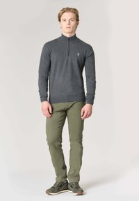 Maglione a collo con zip di colore grigio scuro con maniche lunghe, indossato sopra pantaloni verde chiaro a taglio aderente, abbinato a scarpe da ginnastica scure con un accento beige.