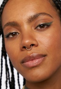 Närbild av ett ansikte med mörk flätad hår, en guldfärgad septumring och grön eyeliner. Huden är jämn med en naturlig läppfärg.