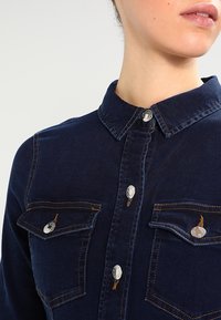 Kvinna som bär en mörkblå denimskjorta med knappade bröstfickor och krage, visad från halsen till midjan.