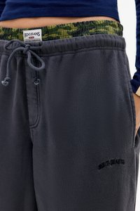 Grå joggingbyxor med en texturerad tyg, som har en elastisk midja med ett grönt camouflage-mönster och en dragsko. "BDG Jeans"-logotypen visas.