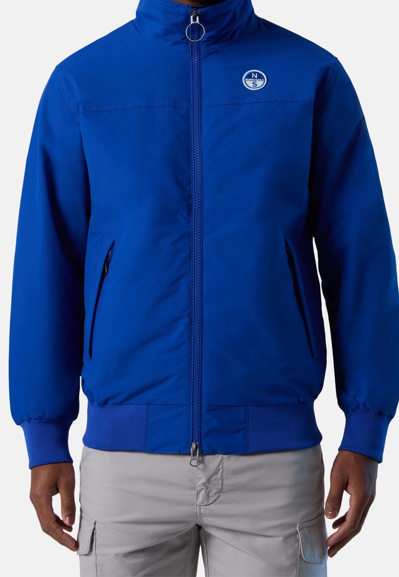Homme portant une veste zippée bleu vif avec un col montant et un logo sur la poitrine gauche, associée à un pantalon cargo gris clair.