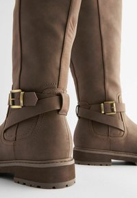 Bottes hauteur genou en daim marron avec semelles épaisses et sangles en cuir décoratives ornées de boucles dorées sur fond blanc.