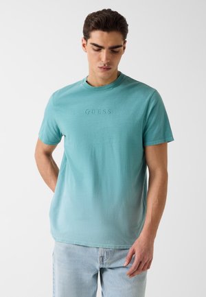 Junger Mann, der ein türkisfarbenes GUESS-T-Shirt und hellblaue Jeans trägt, schaut nach unten, eine Hand hinter dem Rücken, vor einem schlichten weißen Hintergrund.