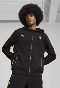 Sort zip-up hættetrøje lavet af blødt stof, med Puma-logo, Ferrari-emblem, sidelommer og kontrastfarvede hvide detaljer langs ærmerne.