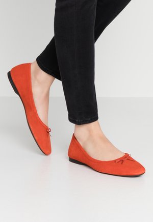 Røde flade ballerinaer i ruskind med en rund tå og en lille sløjfedetail på forfoden, placeret mod sorte jeans på en hvid overflade.