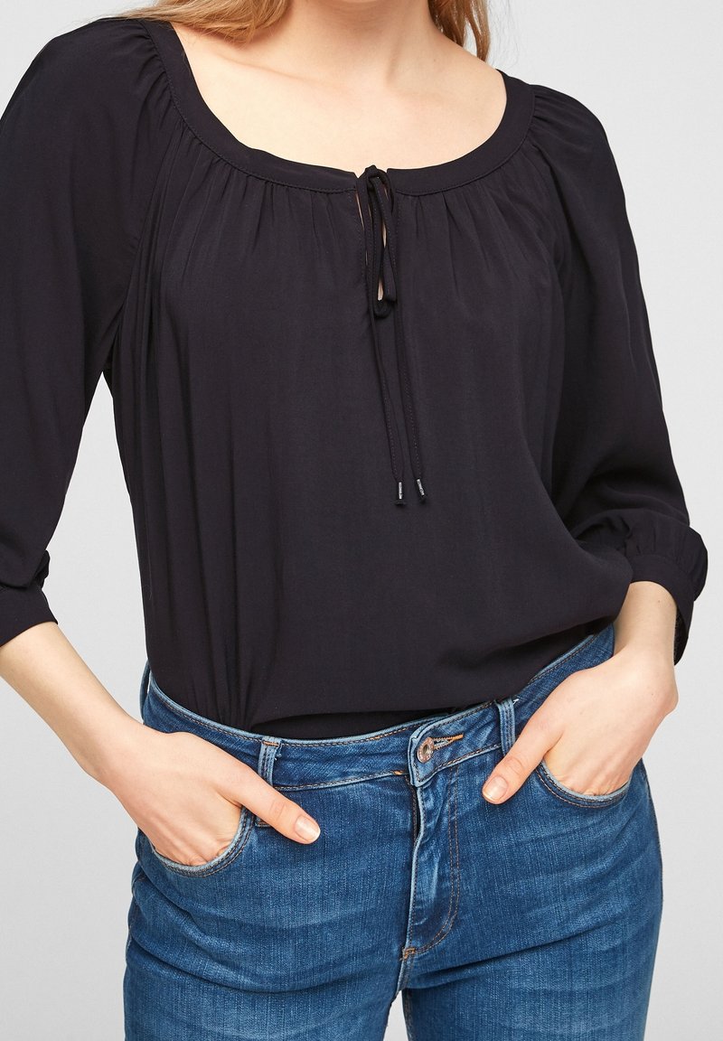 Femme portant un chemisier noir avec un col à nouer et des manches trois-quarts, associé à un jean en denim bleu, mains dans les poches.