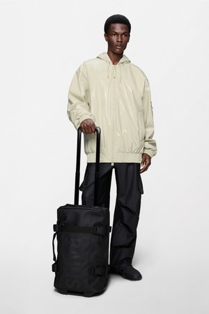 Bolsa de viaje negra con ruedas, correas ajustables, textura suave y detalle de logo. El modelo lleva una chaqueta beige brillante y pantalones negros.