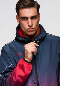 Mladý muž s krátkými hnědými vlasy zapínající námořnickou a červenou větrovku s kapucí, vážný výraz, text na hrudi zní "JEDINÁ HRANICE JSI TY SÁM".