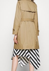 Beżowy płaszcz trencz z paskiem w talii, pagonami na ramionach i klapą z tyłu. Pod spodem asymetryczna spódnica w czarno-białe paski.