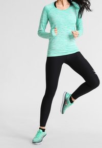 Mintgrünes Sportoberteil mit langen Ärmeln aus strukturiertem Stoff, kombiniert mit schwarzen Leggings und gemusterten mintgrünen Sportschuhen.