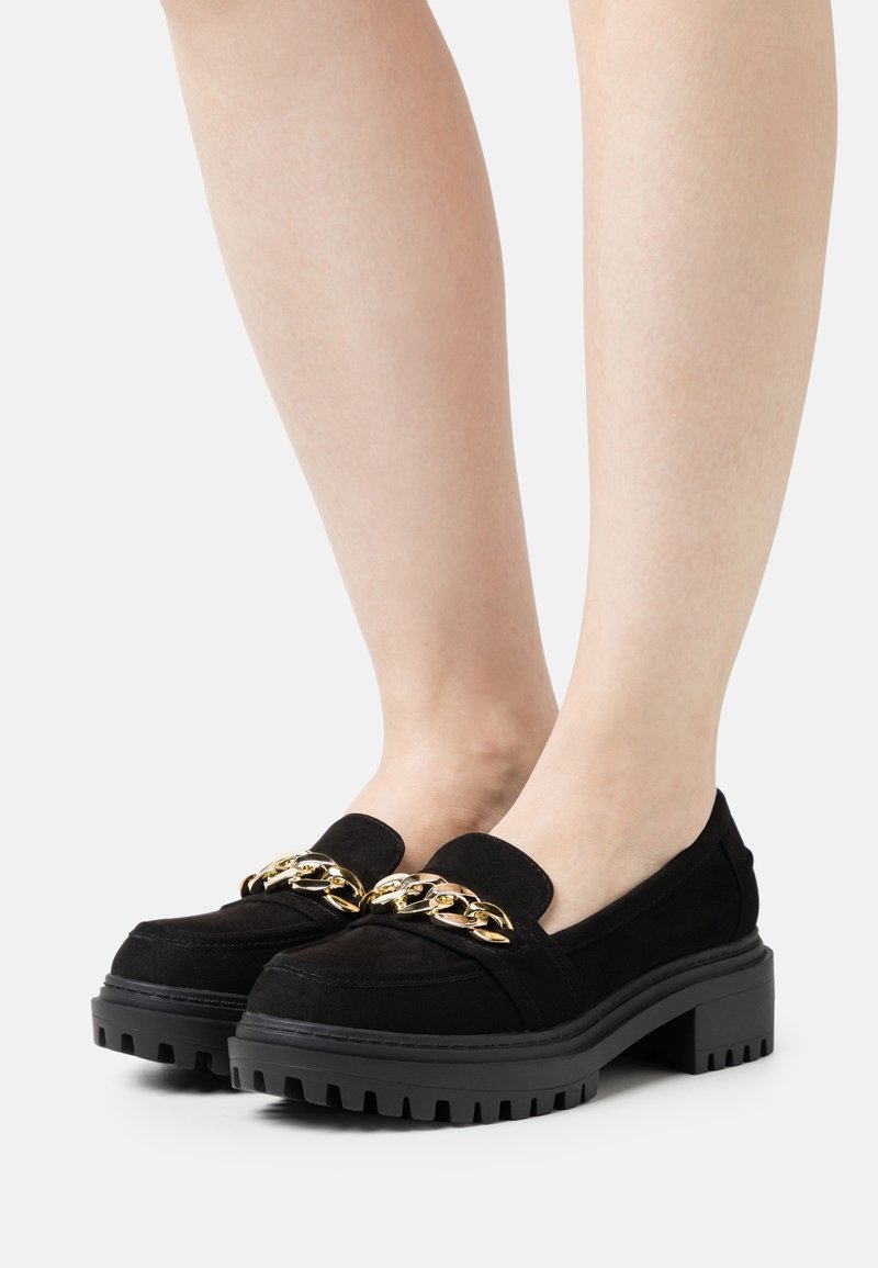 Mocassins en daim noir avec une épaisse semelle en caoutchouc noir, dotés d'un accent en chaîne dorée sur le dessus. Design discret avec un bout arrondi.