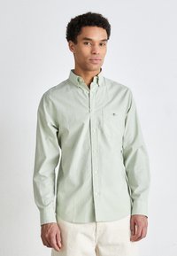 Man wearing light green button-up shirt and beige pants, standing against white background, facing front with neutral expression.