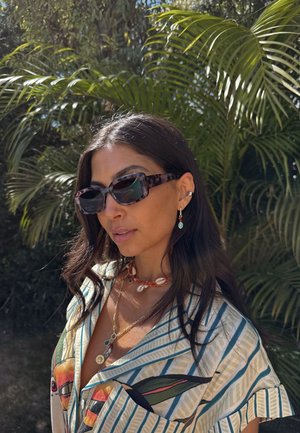 Occhiali da sole tartarugati con forma angolare, catene sovrapposte con conchiglie e perline, e una camicia a righe con motivo. Vegetazione lussureggiante sullo sfondo.