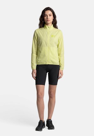 Femme debout portant une veste zippée jaune clair, un short noir au genou et des chaussures de sport noires sur un fond blanc uni.